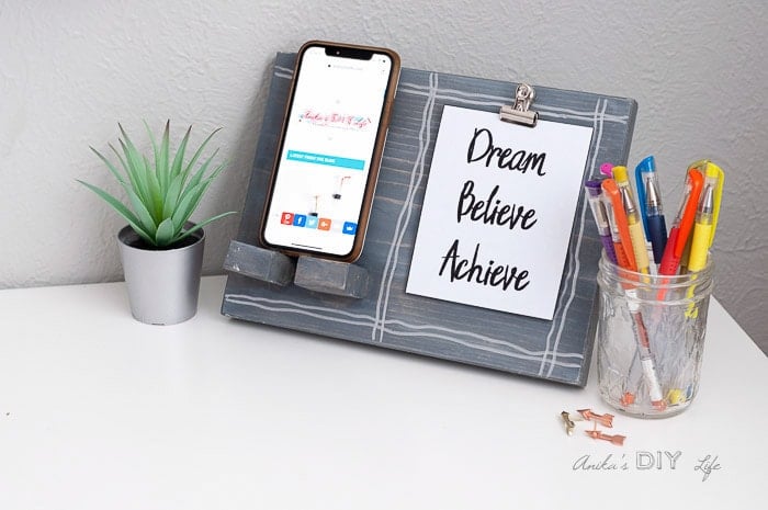DIY phone stand in gray with phone charging, an inspirational quote, pens and a little plant on a desk