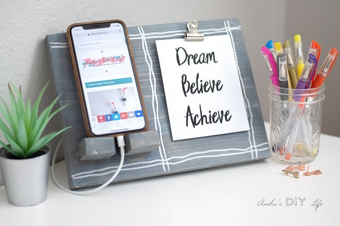 DIY phone stand in gray with phone charging, an inspirational quote, pens and a little plant on a desk