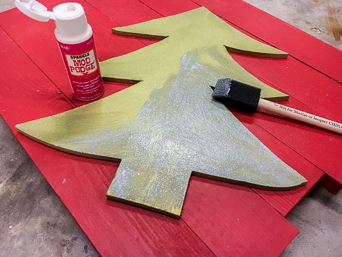 green painted Christmas tree cut out with sparkle mod podge being added.
