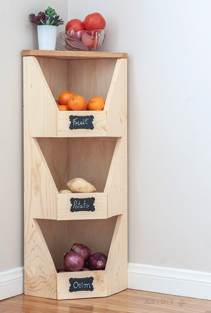 Full DIY corner vegetable storage bin with onions, potatoes and oranges plus bowl of tomatoes on the top.