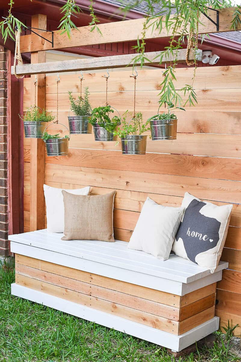 DIY bench outdoors with hanging plants above and pillows on the bench seat