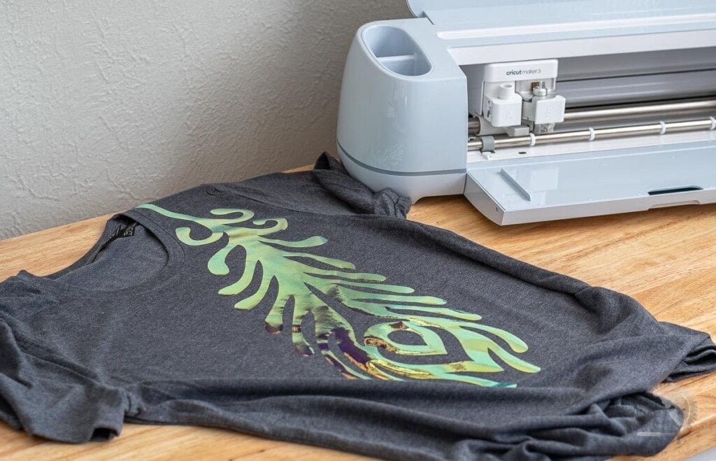 Cricut Maker 3 on table next to gray shirt with holographic feather on it.