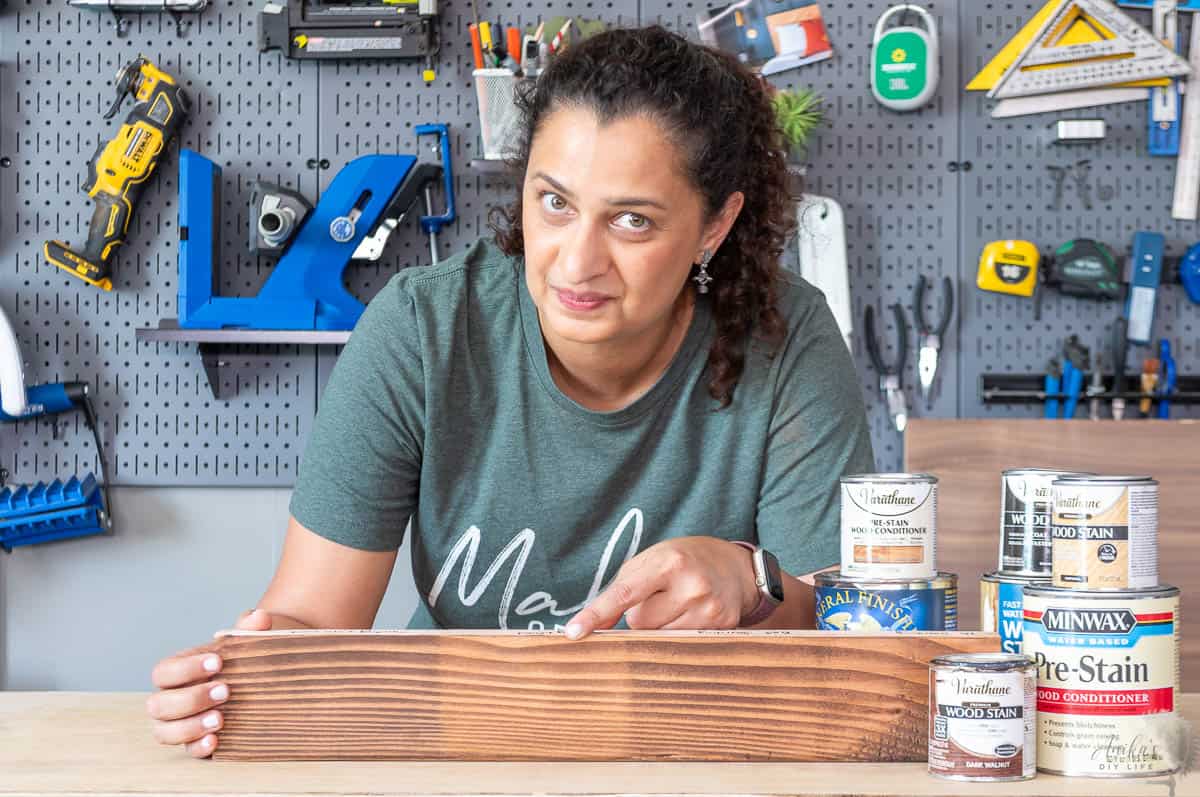 Woman in workshop with a stained 2x4 board showing effects of various conditions on staining 