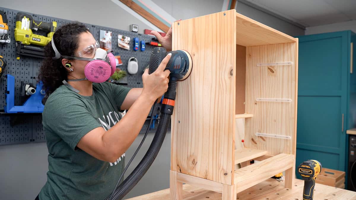 Woman sanding the dresser using a random orbital sander with a face mask on