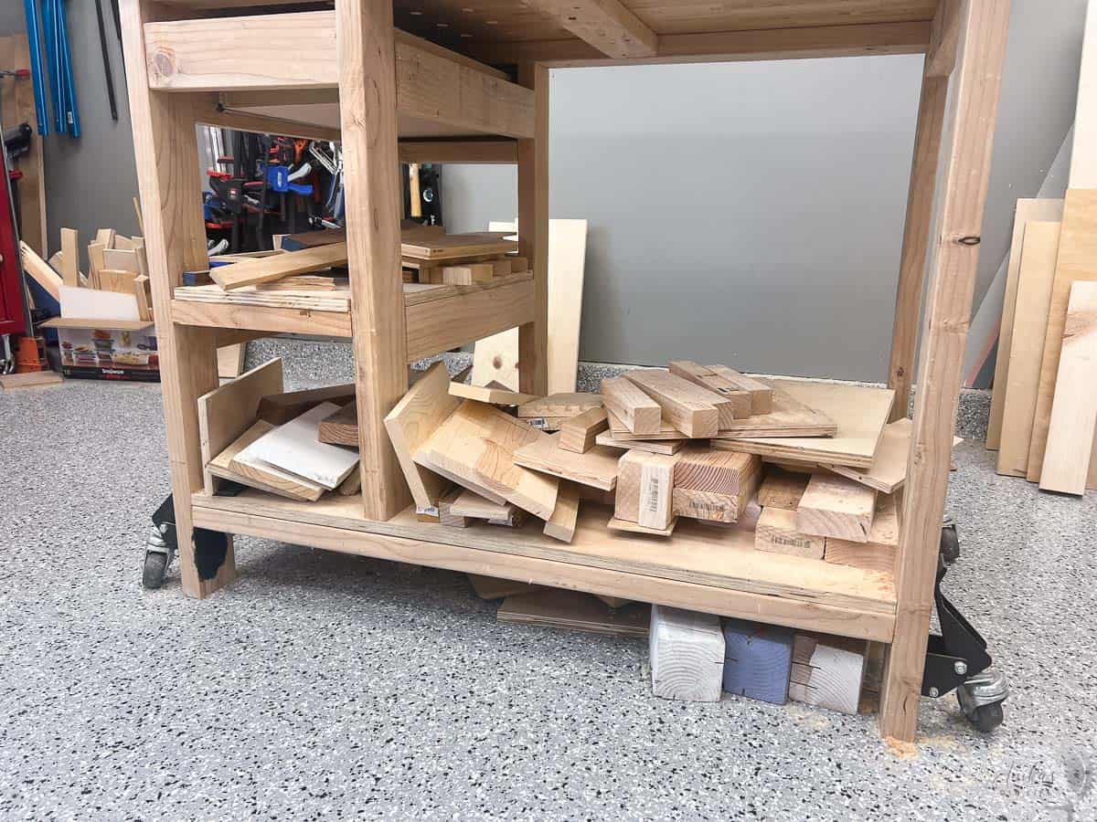 The bottom shelf of a workbench with lots of scrap wood in a messy pile in a workshop