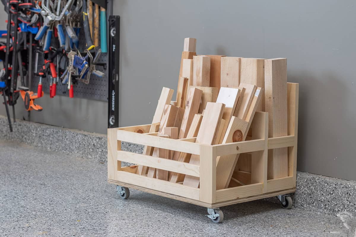A wooden cart on wheels is filled with various sizes of wooden planks and scraps. The crate like cart is placed against a gray wall on a speckled floor.