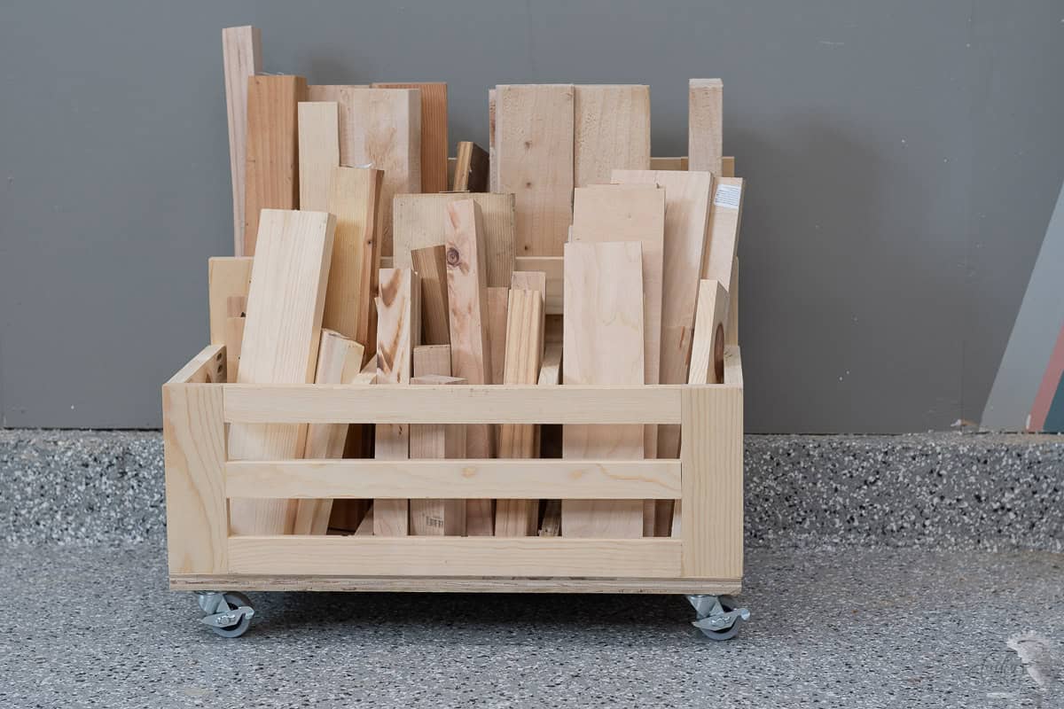 Front view of the diy scrap wood storage cart filled with various sizes of wooden planks and scraps. The crate like cart is placed against a gray wall on a speckled floor.