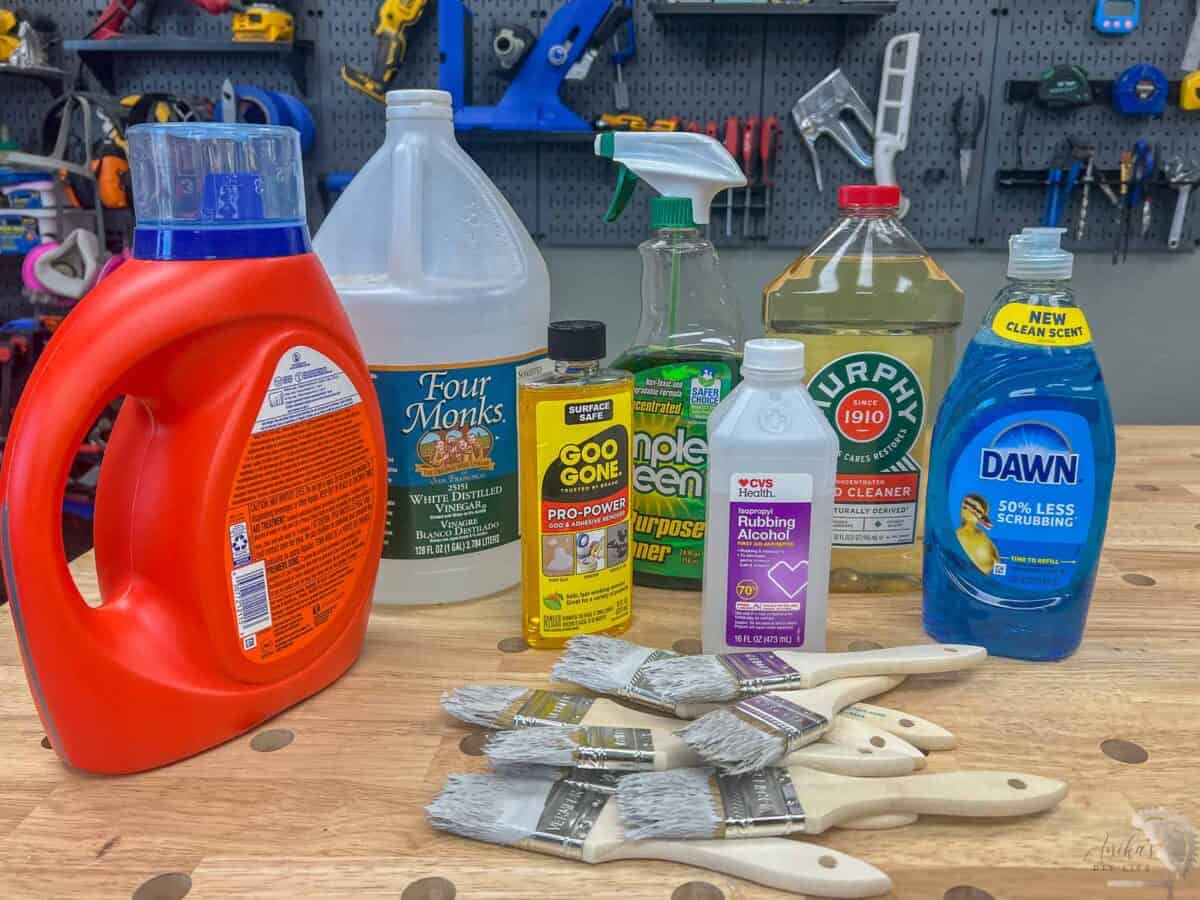 inegar, goo gone, rubbing alcohol, simple green, dawn dish soap, laundry detergent, and murphys oil soap on a workbench with a stack of paint brushes with dried paint.