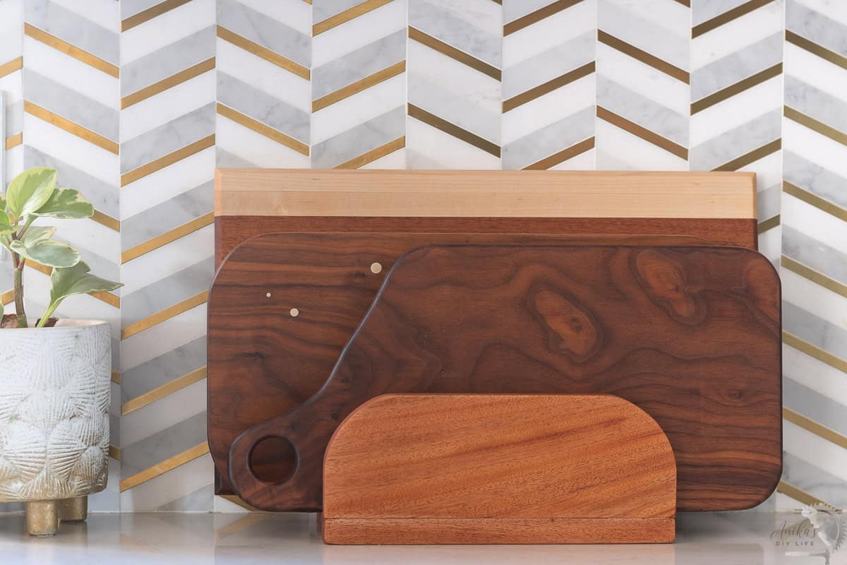 DIY cutting board stand in the kitchen with three walnut cutting boards in it against a gold and grey backsplash.