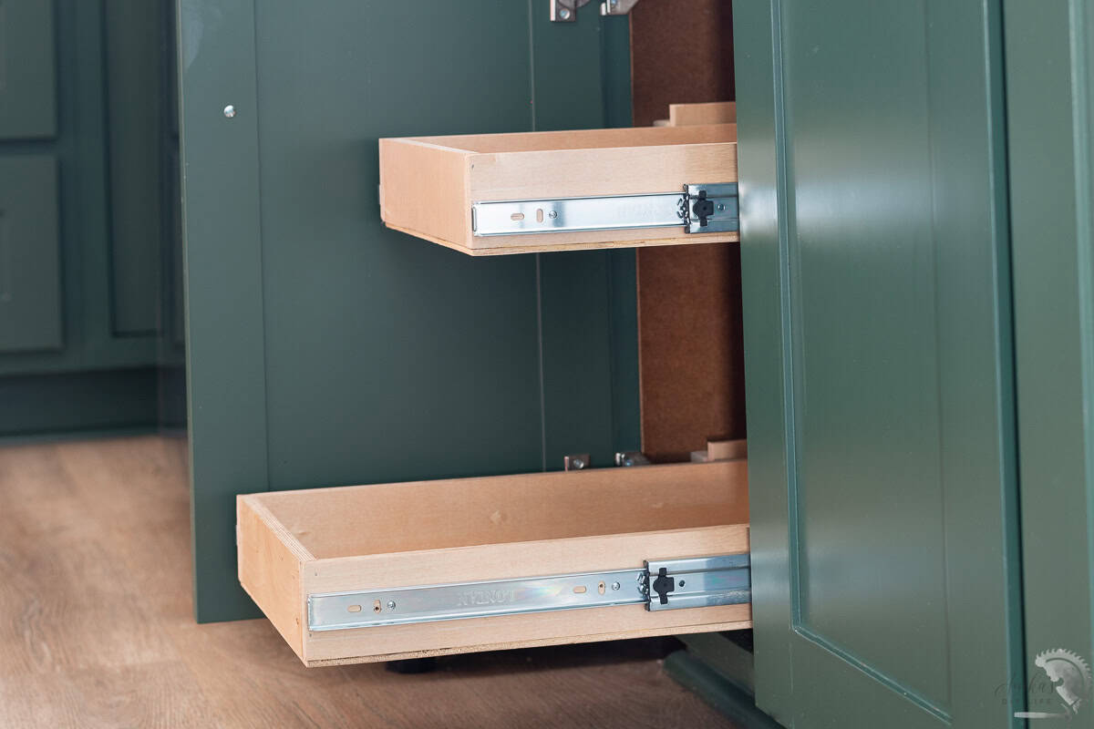 DIY pull-out shelves installed inside a deep green kitchen cabinet. The wooden sliding drawers are mounted on metal drawer slides, making storage more accessible and organized.