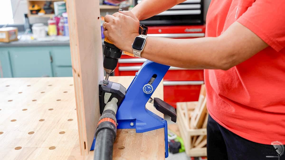 Drilling pocket holes into a board using a blue Kreg 720 pocket hole jig with an attached shop vac for dust collection.