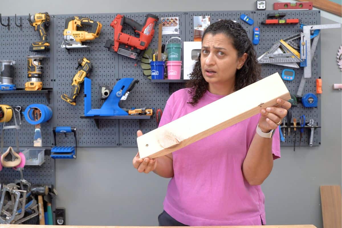 Anika holding a 2x4 with a bewildered expression in a workshop with tools in the background. 
